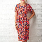A woman in a colorful, floral dress stands against a white brick wall. Her hand on her hip adds a confident, cheerful tone.