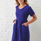 A woman wearing a vibrant blue dress smiles with hands in pockets, standing against a textured white brick wall. The mood is cheerful and stylish.
