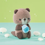 Cute crocheted otter holding a textured blue seashell sits on a teal surface with scattered white seashells, set against a light green background.