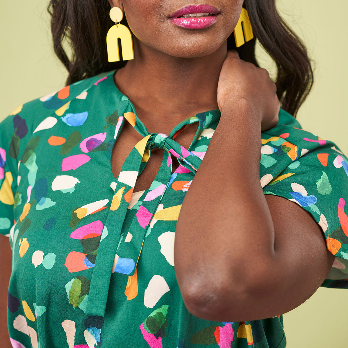 A woman wears a vibrant green dress with colorful abstract patterns and bold yellow earrings. Her expression is serene with a soft, relaxed posture.