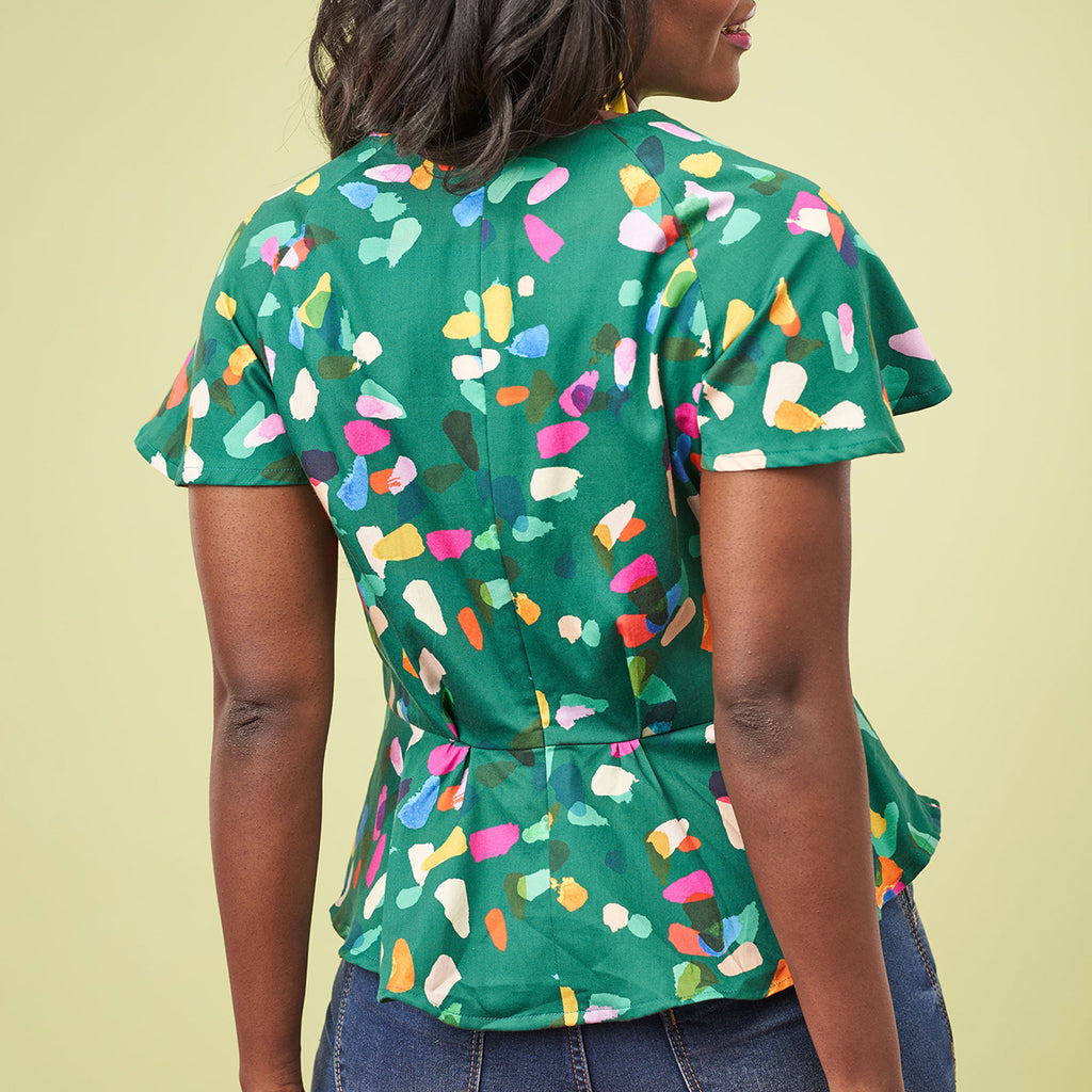 A woman with shoulder-length hair is seen from behind wearing a vibrant green blouse with colorful abstract patterns, paired with denim jeans.