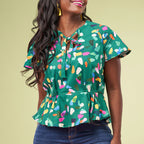 A woman wears a colorful, abstract-patterned green blouse with short sleeves and a tie at the neckline. She smiles joyfully against a light green background.