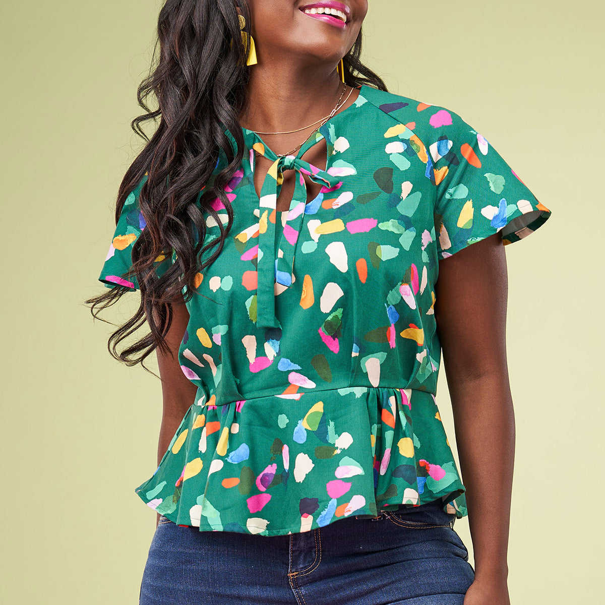 A woman wears a colorful, abstract-patterned green blouse with short sleeves and a tie at the neckline. She smiles joyfully against a light green background.