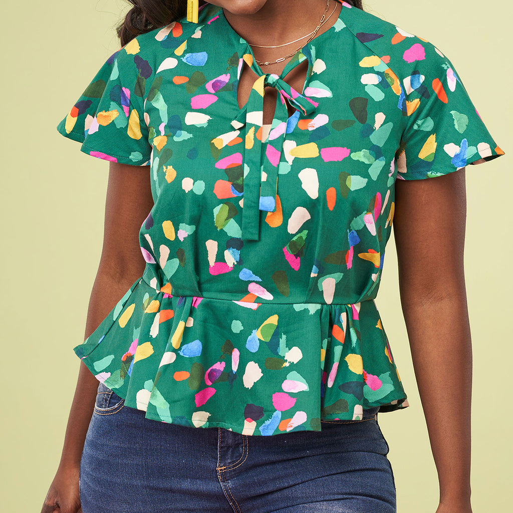 Woman in colorful abstract-patterned green blouse with short sleeves and a tie at the neckline. She is wearing dark jeans. The tone is vibrant and cheerful.