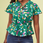 Woman in colorful abstract-patterned green blouse with short sleeves and a tie at the neckline. She is wearing dark jeans. The tone is vibrant and cheerful.