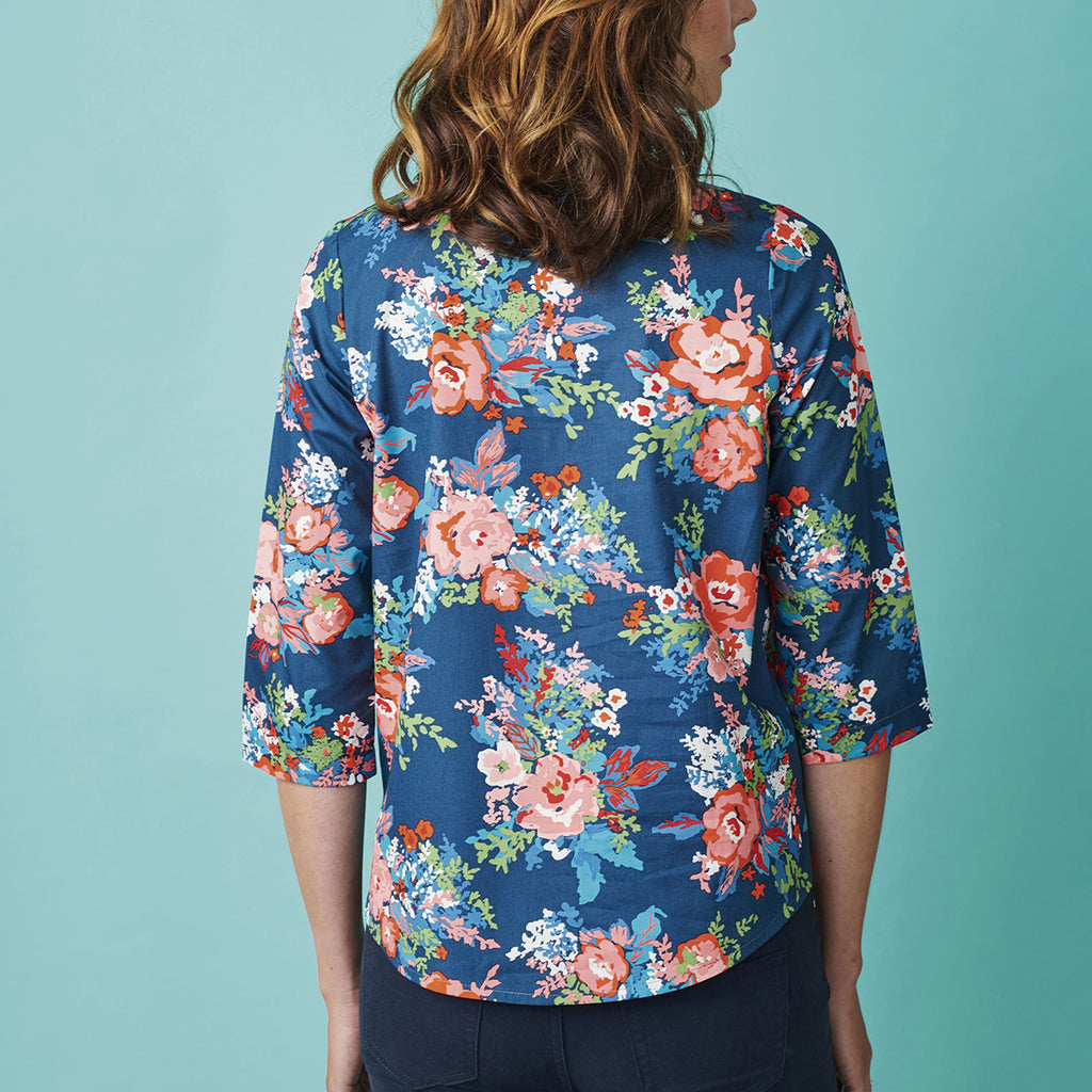 Woman with wavy hair, facing away, wears a vibrant blue blouse with pink floral patterns. Teal background, casual and colorful mood.