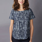A woman in a short-sleeved, patterned blouse stands against a gray background, smiling warmly. The blue blouse has a linear, abstract design.