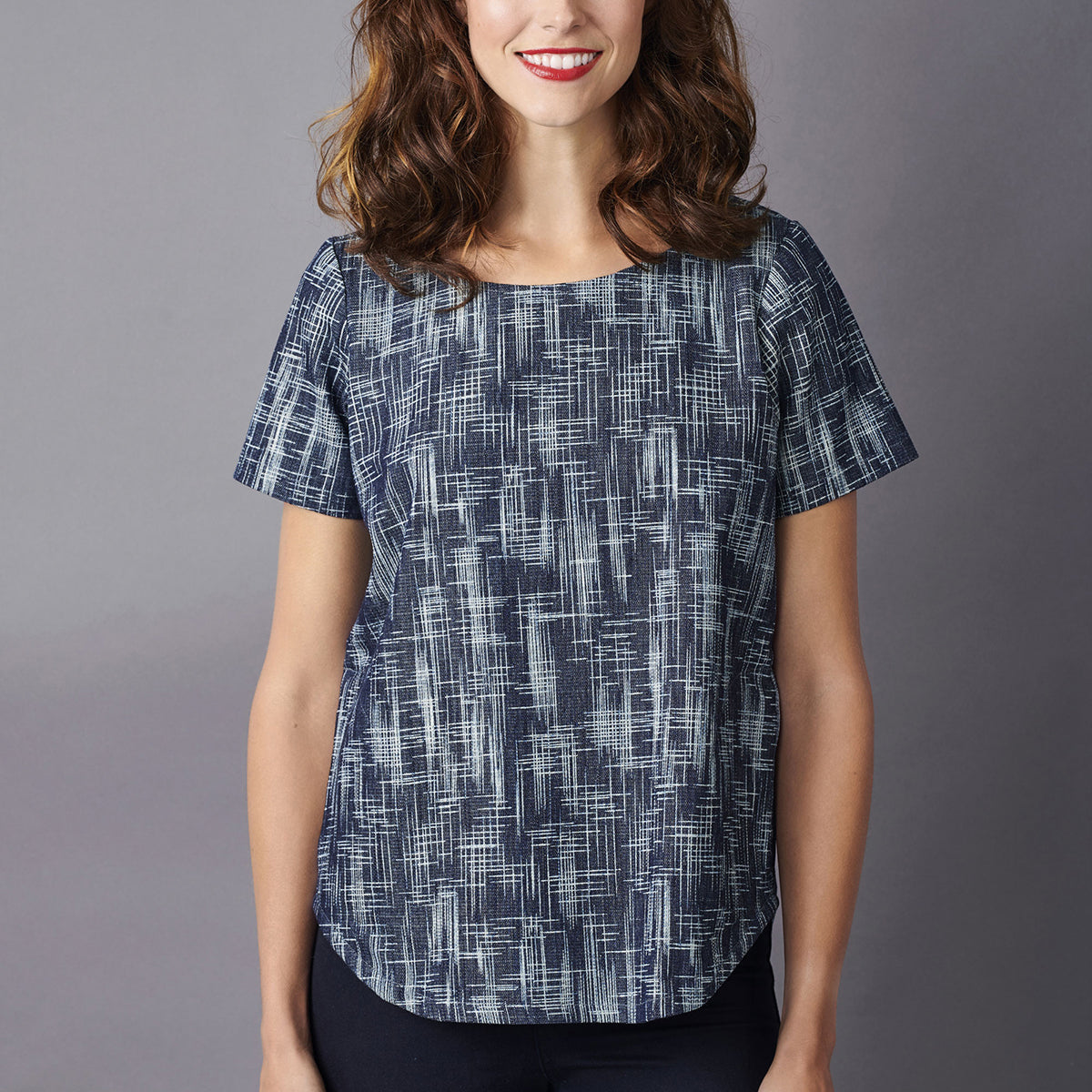 A woman in a short-sleeved, patterned blouse stands against a gray background, smiling warmly. The blue blouse has a linear, abstract design.