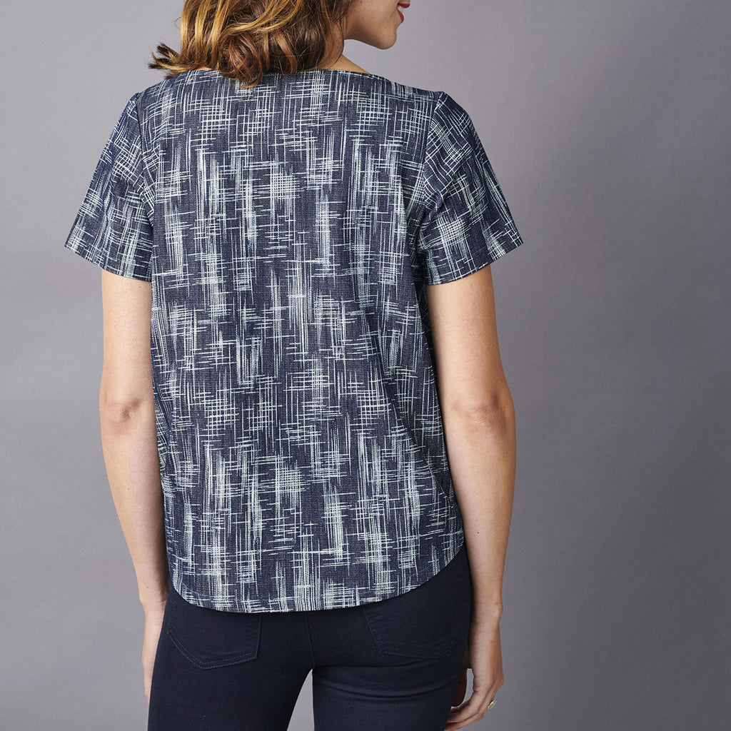 A person with wavy brown hair is facing away, wearing a short-sleeved blouse with a navy and white abstract grid pattern, paired with dark jeans.