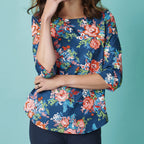 A woman wearing a vibrant floral blouse with shades of red, pink, and blue on a dark background. The mood is bright and cheerful. She stands against a light teal backdrop, her arms folded.