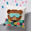 Decorative pillow with a bear face and sleep mask, surrounded by colorful 'Z' letters on a light gray background.