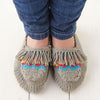 A pair of knitted moccasin slippers with multicolored tassels on the toes, worn by a person sitting against a white background.