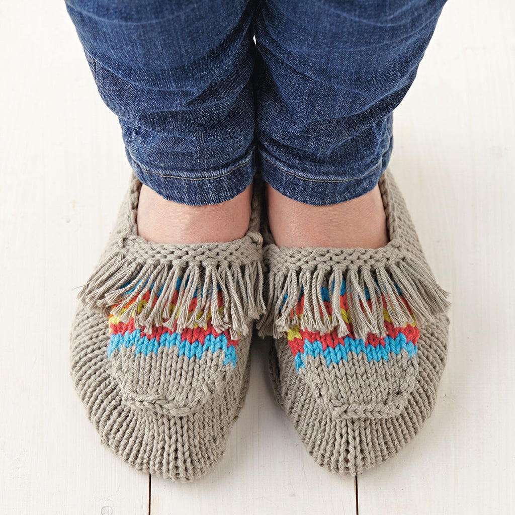 A pair of knitted moccasin slippers with multicolored tassels on the toes, worn by a person sitting against a white background.