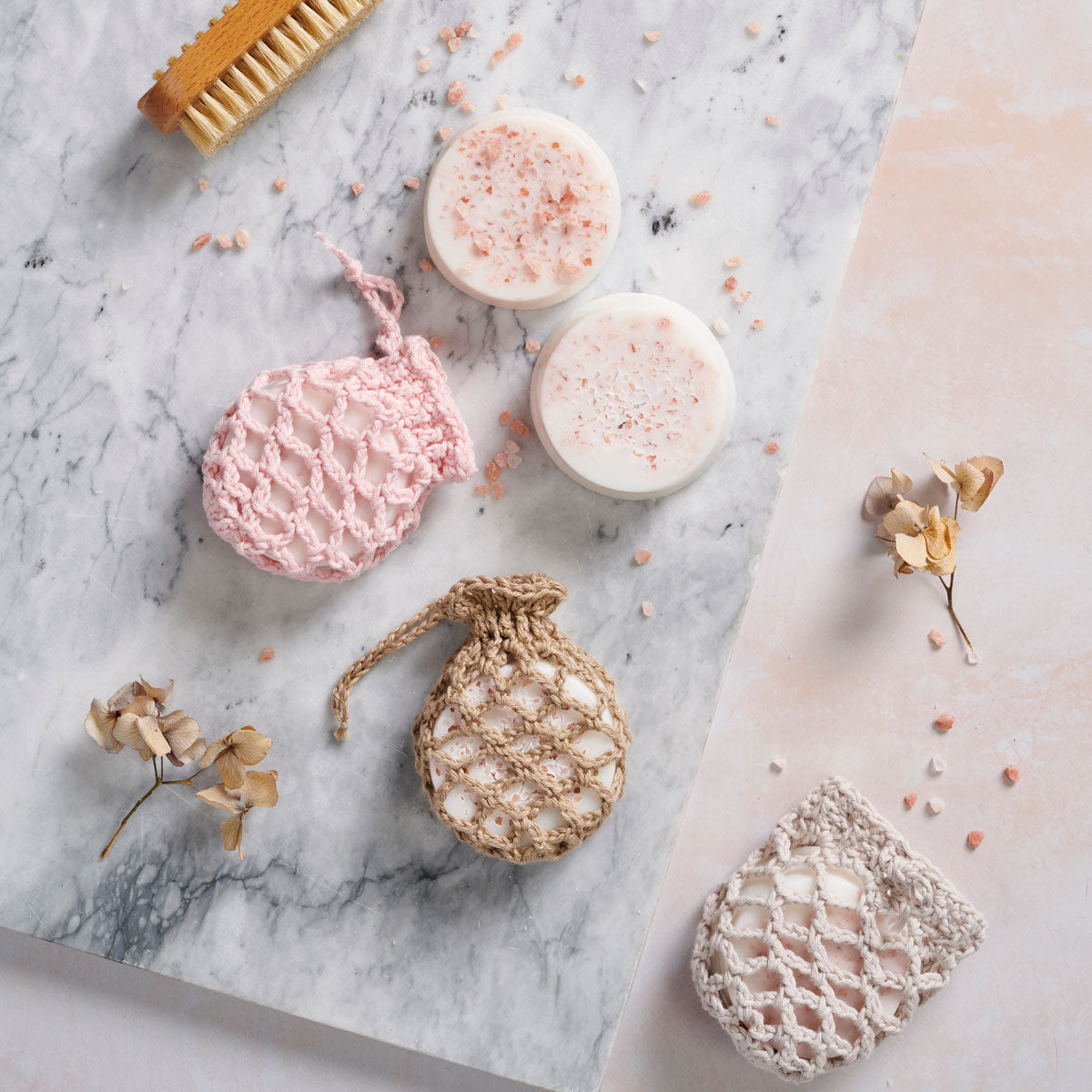 A collection of crochet patterns for soap pouches alongside a bar of soap with scattered Himalayan pink rock salt, displayed on a marble surface with a small brush and decorative flowers.