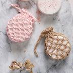 Two handmade soaps in crocheted pouches on a marble surface. One pink, one beige, with Himalayan salt. Accented by dried flowers. Calm and natural vibe.