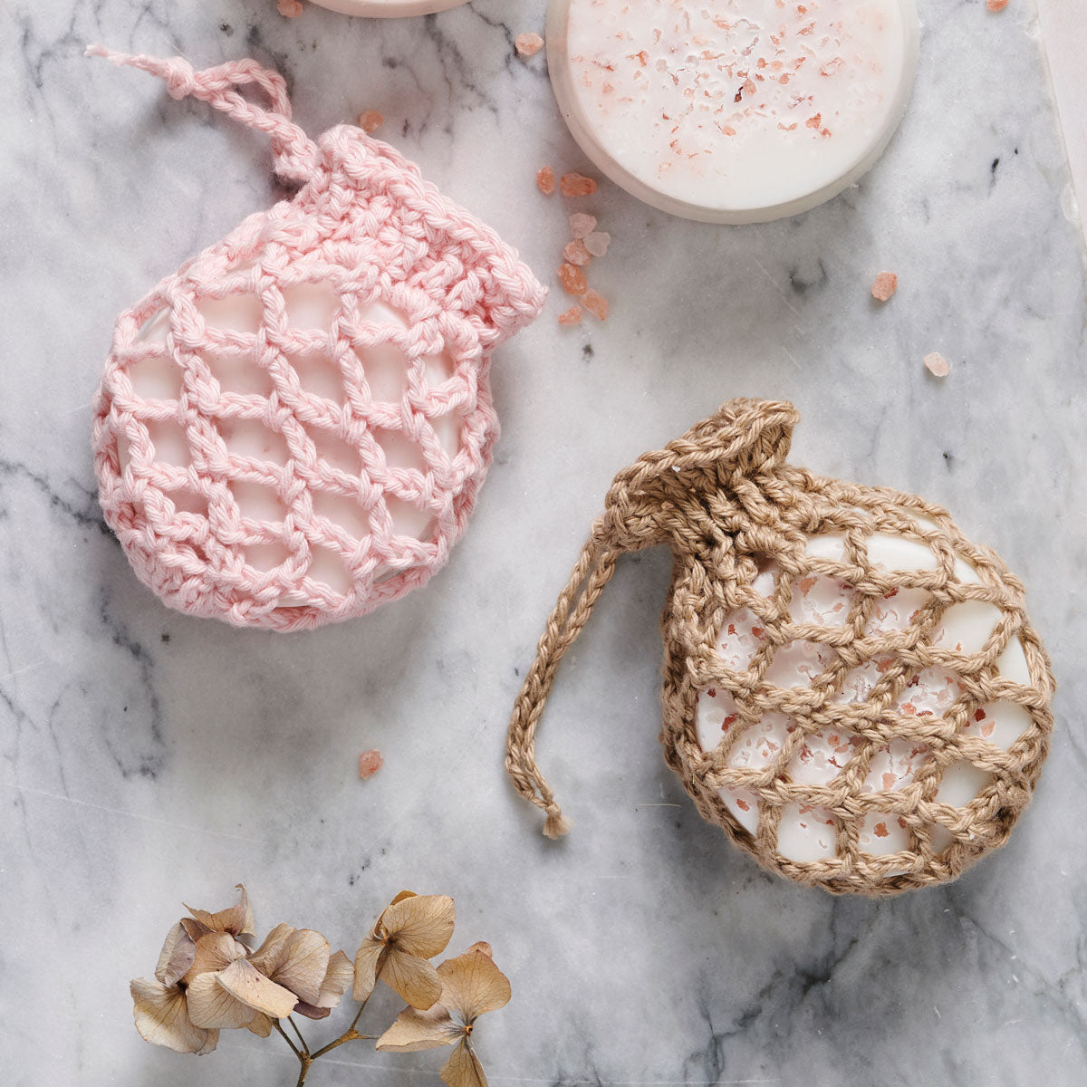 Two handmade soaps in crocheted pouches on a marble surface. One pink, one beige, with Himalayan salt. Accented by dried flowers. Calm and natural vibe.