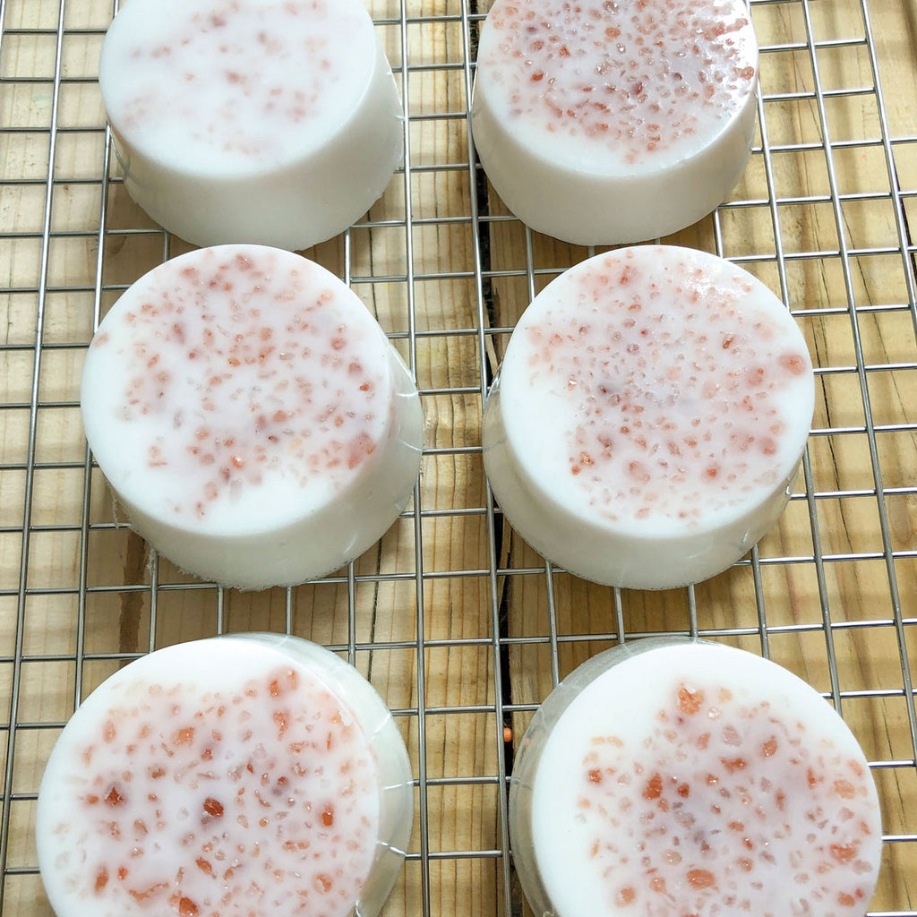 Round soap bars with white and pink marbled patterns are cooling on a wire rack. The smooth texture and gentle colors suggest freshness and care.