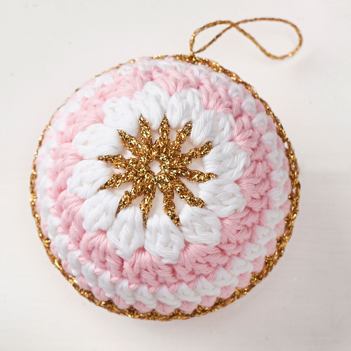 A crochet ornament featuring a white and pink spiral pattern, accented with a central gold star and edge. It hangs by a thin gold loop, exuding a festive feel.