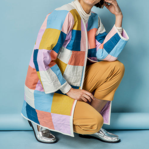 Person wearing a colorful patchwork coat and brown pants on a blue background