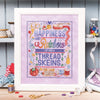 Framed cross-stitched artwork with colorful text and sewing-themed elements on a wooden surface.