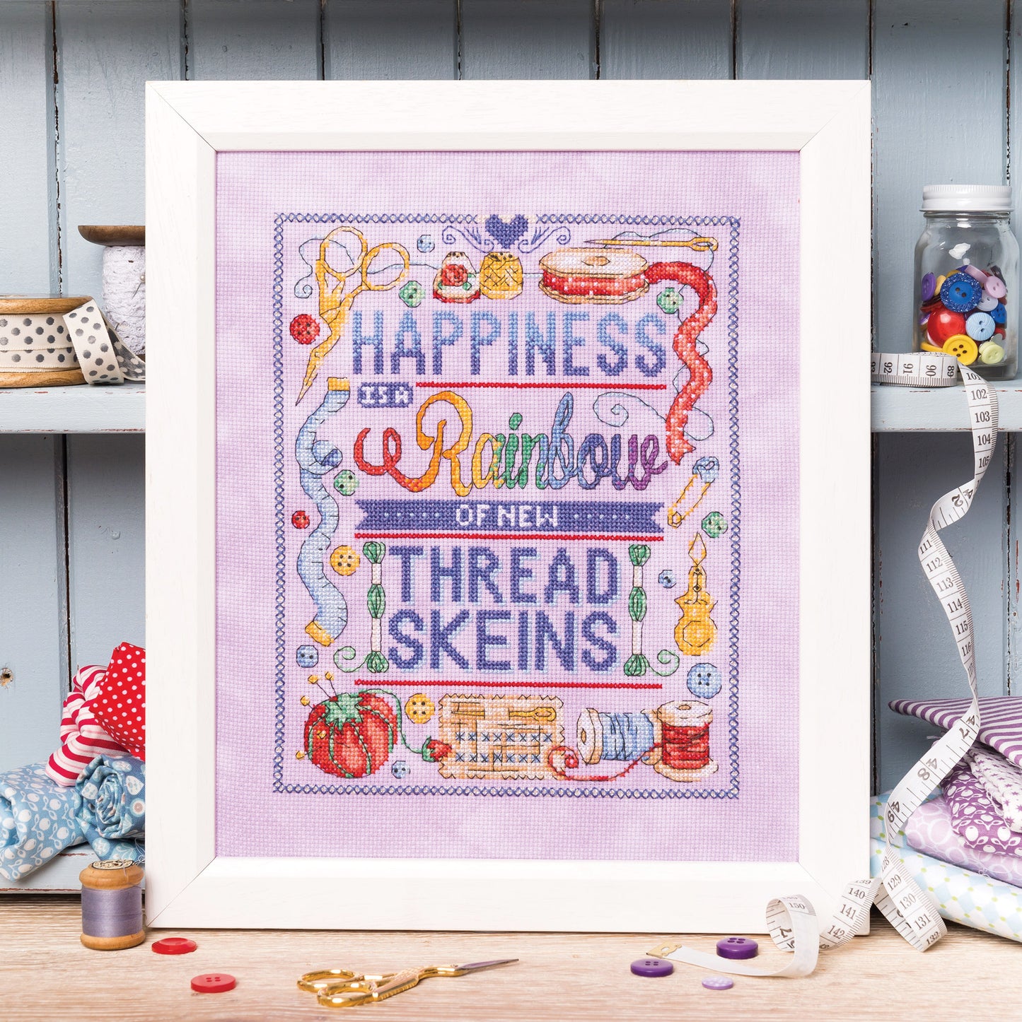 Framed cross-stitched artwork with colorful text and sewing-themed elements on a wooden surface.
