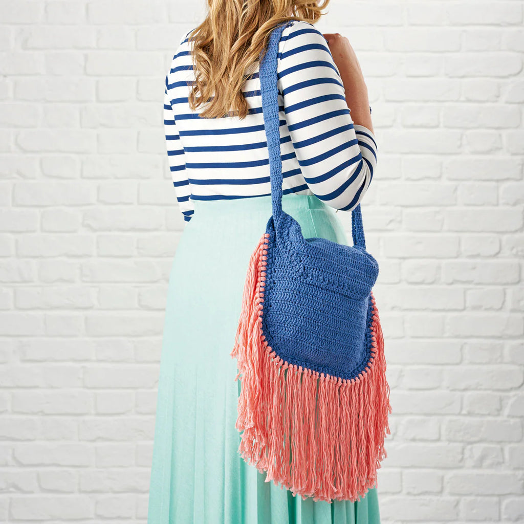 Woman holding a blue and pink fringed bag against a white brick wall.