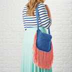 Woman holding a blue and pink fringed bag against a white brick wall.