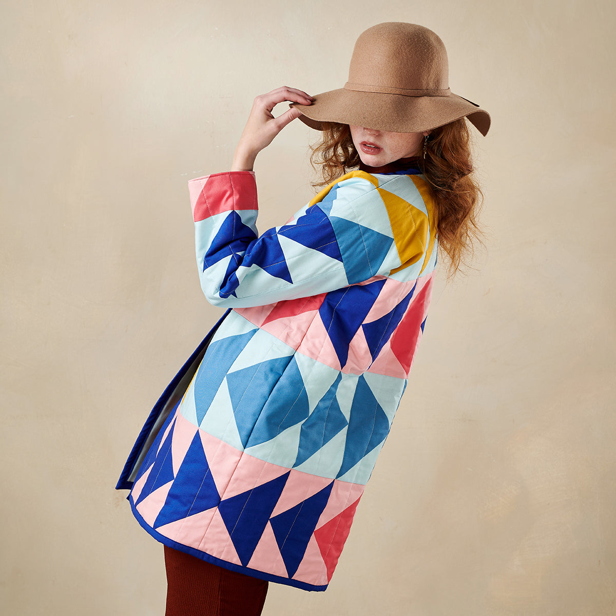 Woman wearing a colorful geometric-patterned coat and brown hat against a beige background