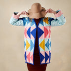 Person wearing a colorful geometric-patterned jacket and a wide-brimmed hat on a beige background