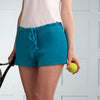 Person wearing teal crochet shorts holding a tennis racket and ball against a light background
