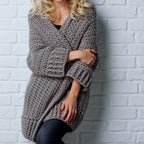 Woman wearing a chunky knit gray sweater against a light gray brick wall.