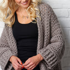 Woman wearing a chunky knit gray cardigan against a white brick wall.