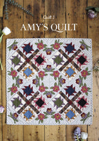 Floral patterned quilt titled "Amy's Quilt" on rustic wooden floor. Features geometric shapes and flowers in red, blue, and green. Elegant, cozy feel.