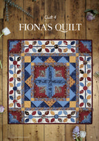 A vibrant quilt named "Fiona's Quilt" on wooden floorboards. The quilt features intricate patterns in blue, red, and yellow with floral motifs.
