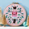 Embroidery hoop with a nutcracker design on a pink background, surrounded by small Christmas trees.