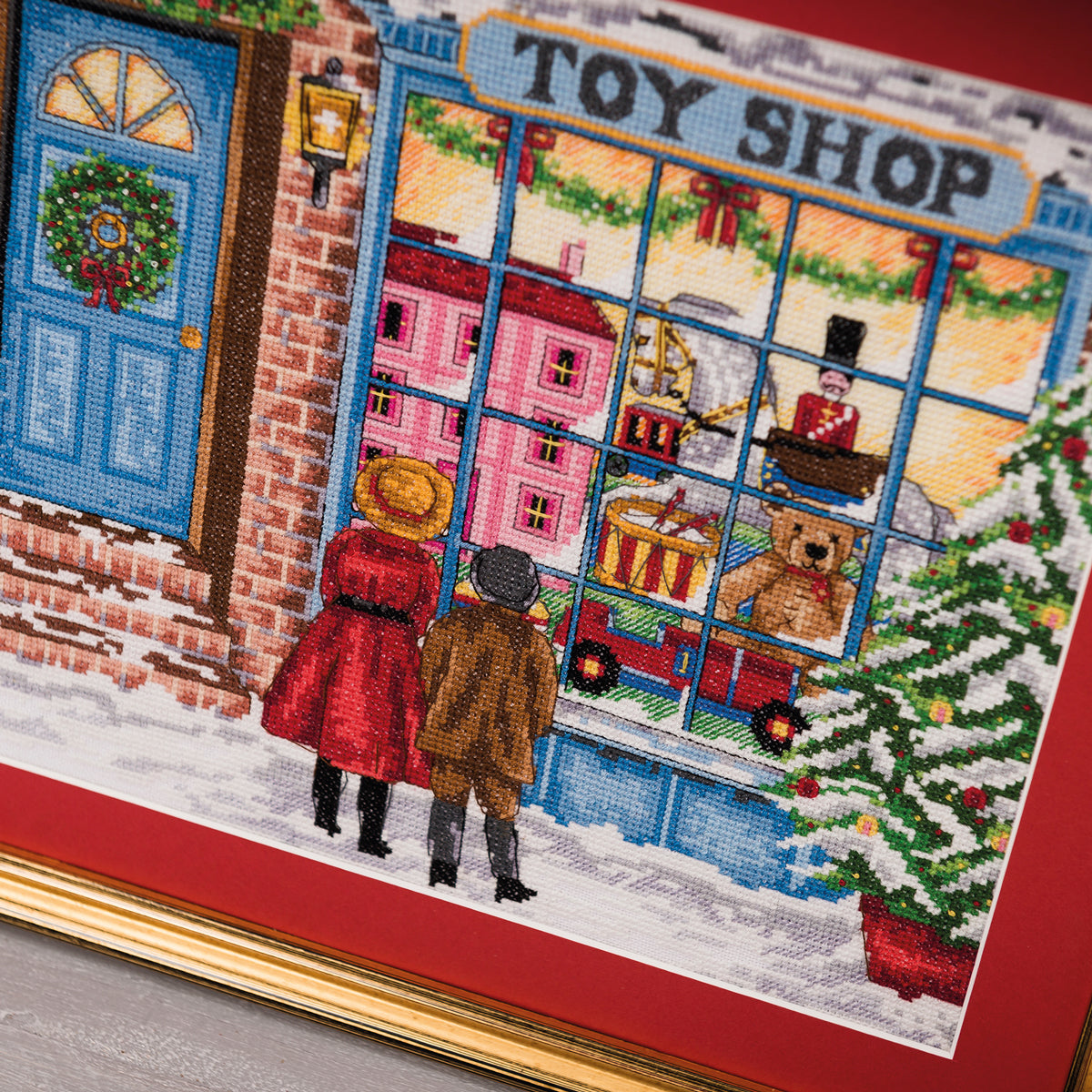 Cross-stitched scene of a toy shop window with two people looking in, Christmas tree on right
