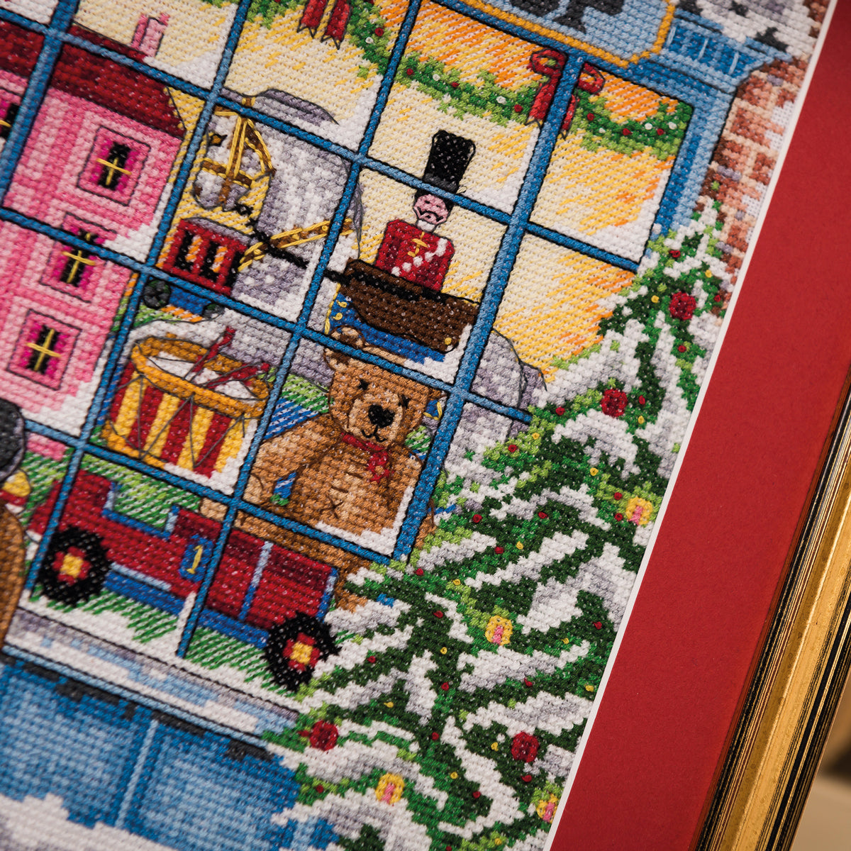 Cross-stitched scene with a teddy bear, Christmas tree, and festive elements on a red background.