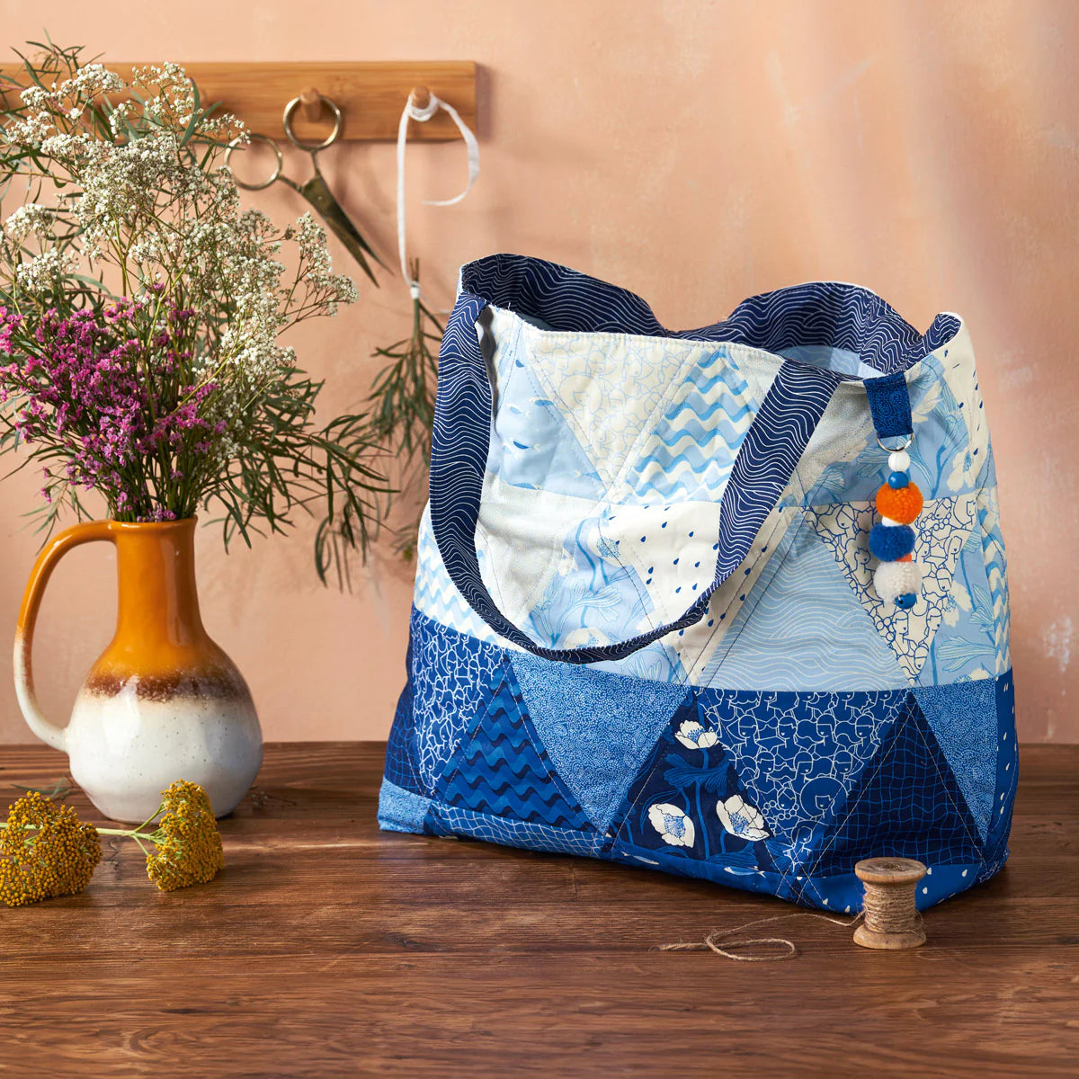 Blue and white patchwork bag on a wooden surface with a vase and flowers in the background.