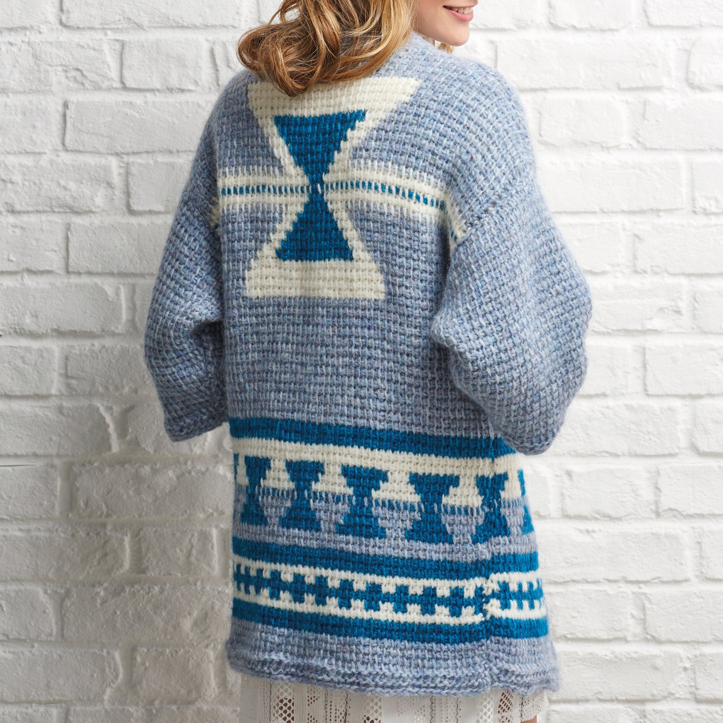Person wearing a blue and white patterned crocheted sweater against a white brick wall.