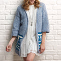 Woman wearing a blue knitted cardigan with patterned sleeves against a white brick wall.