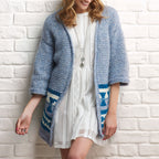 Woman wearing a blue knitted cardigan with patterned sleeves against a white brick wall.