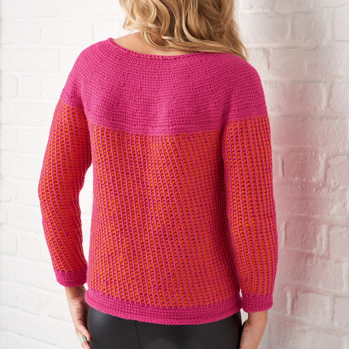 A woman with wavy blonde hair stands facing a white brick wall, wearing a crocheted sweater in vibrant pink and orange tones.
