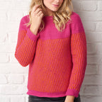 A woman in a bright pink and orange knitted sweater stands against a white brick wall. Her wavy blonde hair cascades over her shoulders as she smiles softly.