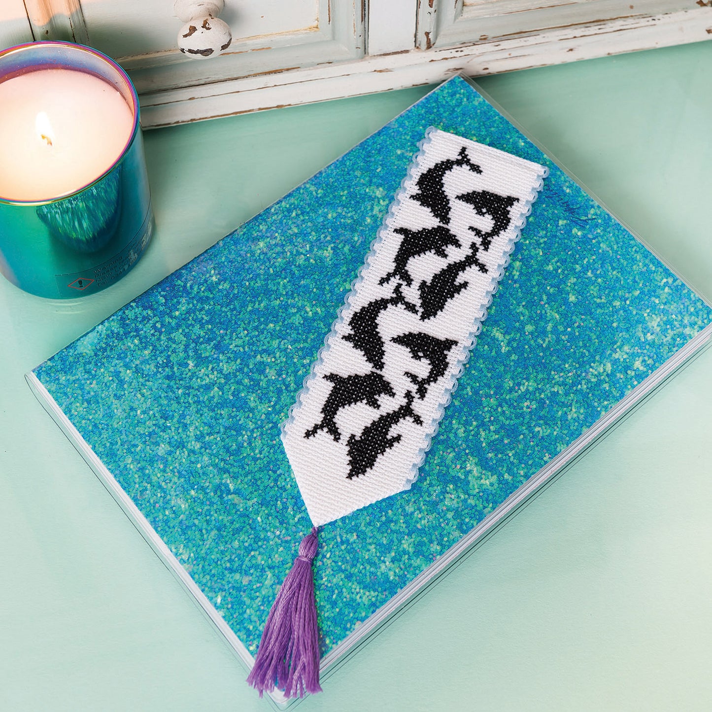 Book with a beaded bookmark featuring dolphins on a teal surface next to a candle.