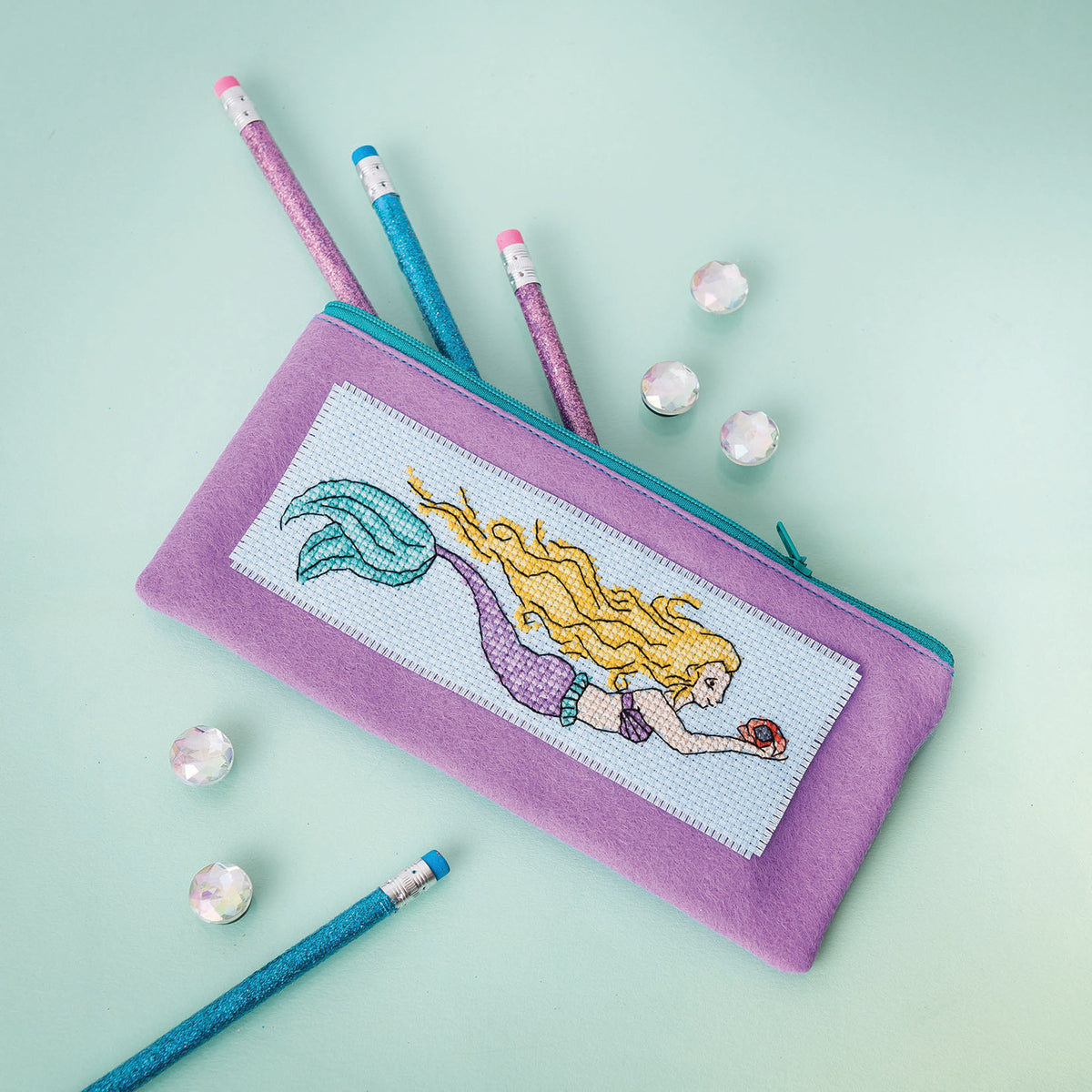 Cross-stitched mermaid design on a pink pencil case with pencils on a light green background