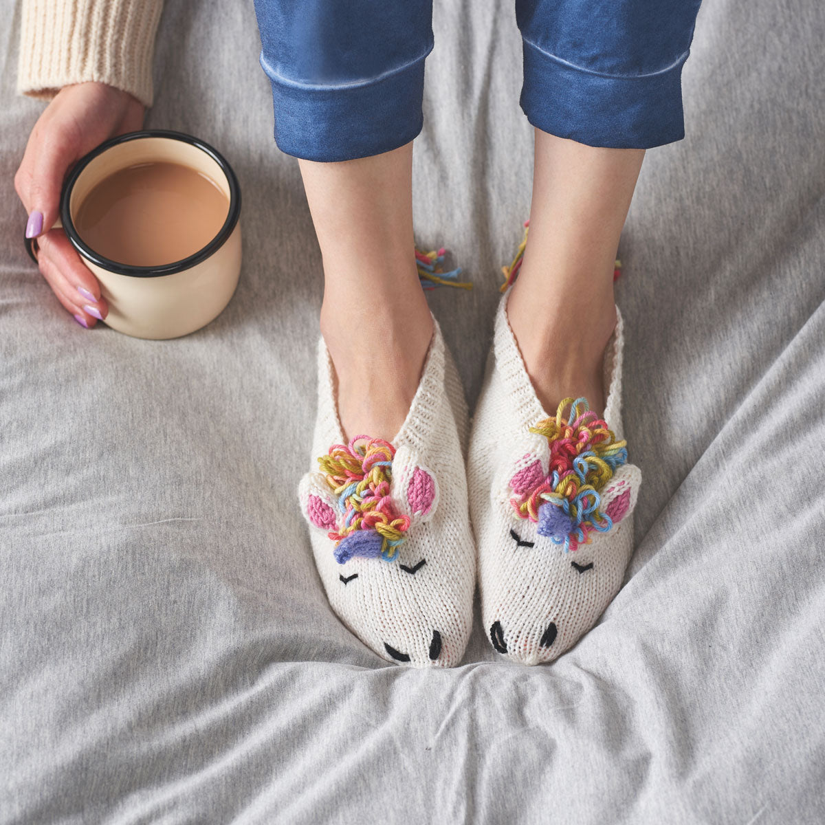 A pair of knitted unicorn slippers with multicolored mane and tail, being worn by a person sitting on a bed with a cup of tea.