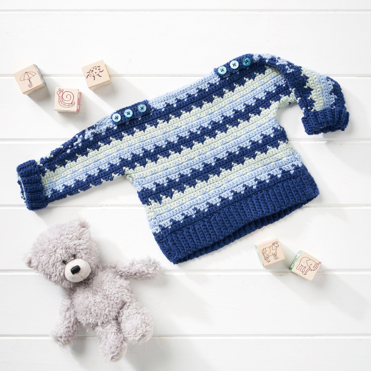 Blue and white striped baby sweater with a teddy bear and wooden blocks on a light wooden surface