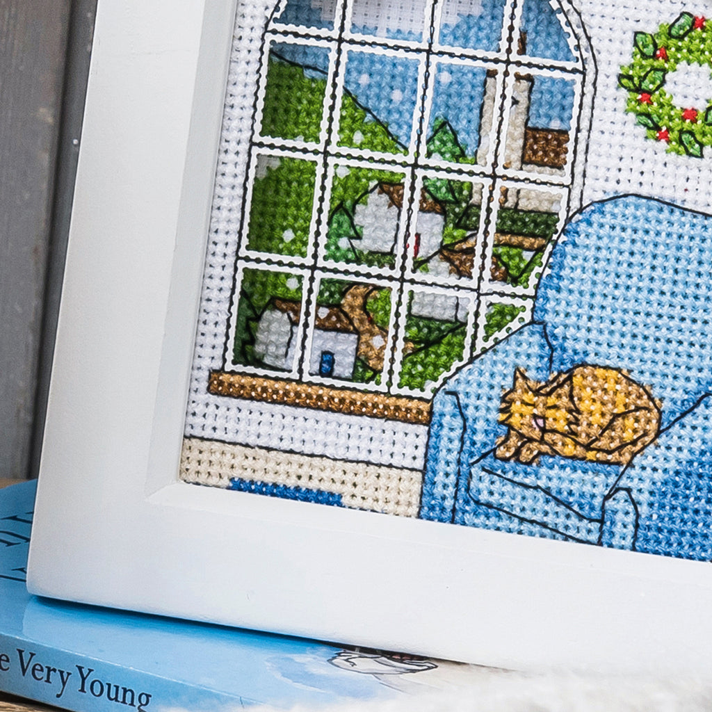 Cross-stitched scene with a cat on a couch and a window view, on top of a book.