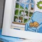 Cross-stitched scene with a cat on a couch and a window view, on top of a book.