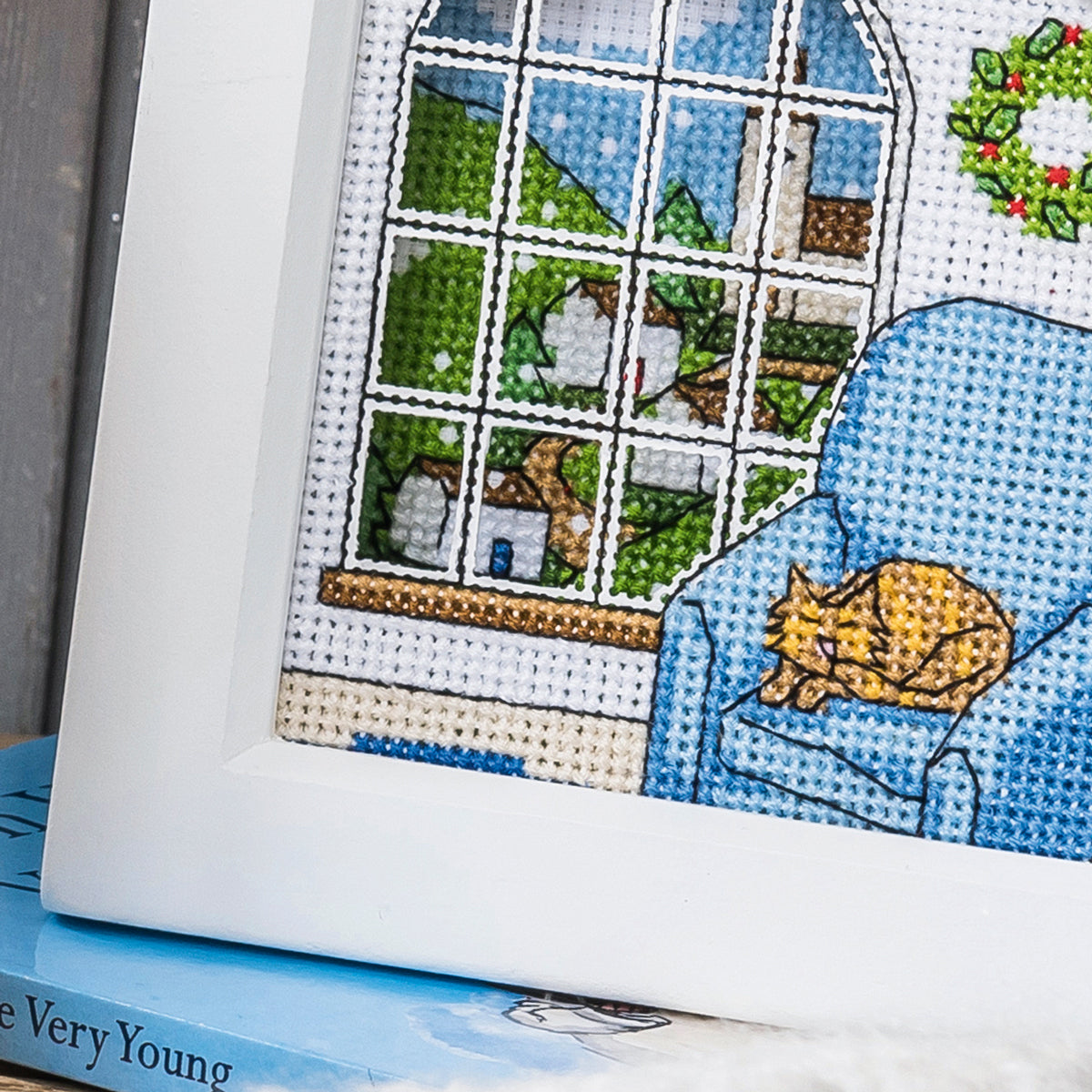 Cross-stitched scene with a cat on a couch and a window view, on top of a book.